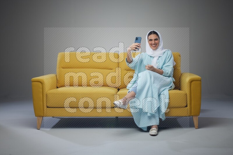A Saudi woman wearing a light blue Abaya and white head scarf sitting on a yellow sofa and taking selfies with her phone eye level on a grey background