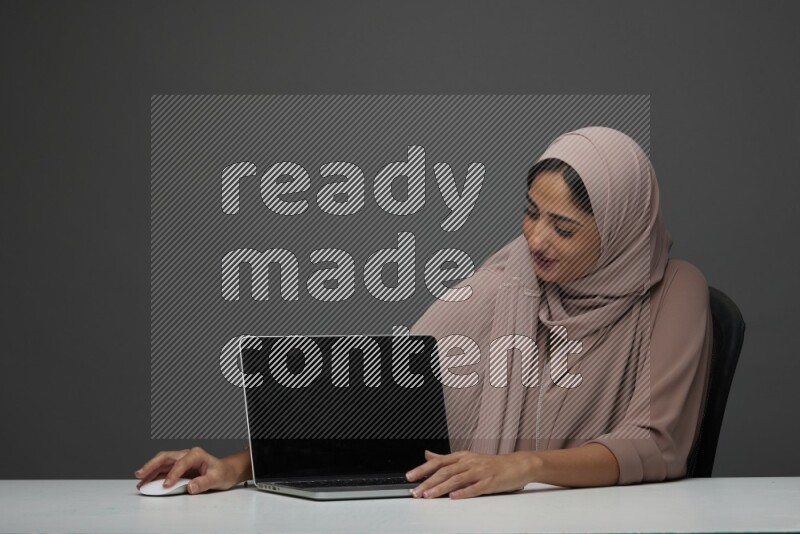A woman Sitting on her desk  Pointing at her laptop on a Gray Background wearing Brown Abaya with Hijab