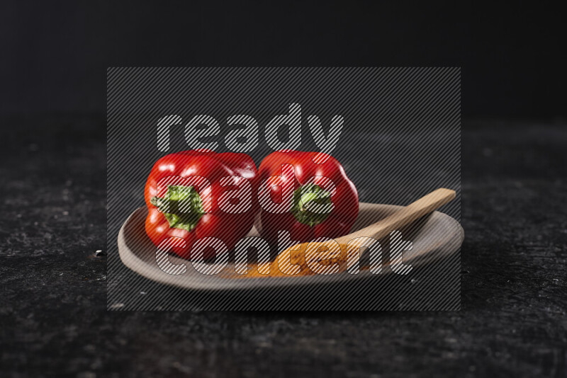 A wooden spoon full of ground paprika powder with red bell peppers, all on a pottery plate on black background