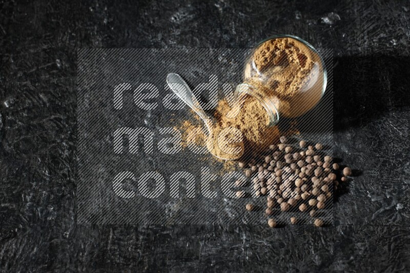 A flipped glass spice jar and metal spoon full of allspice powder and powder spilled out of it with whole balls on a textured black flooring