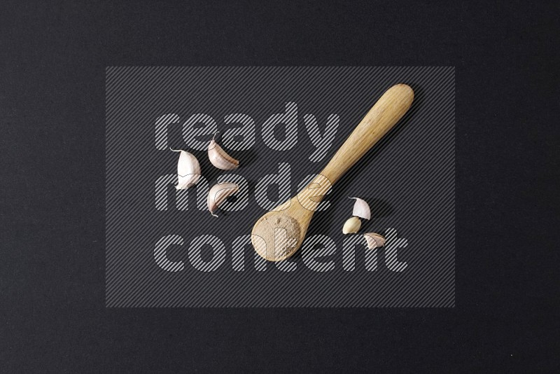 A wooden spoon full of garlic powder with cloves beside it on a black flooring