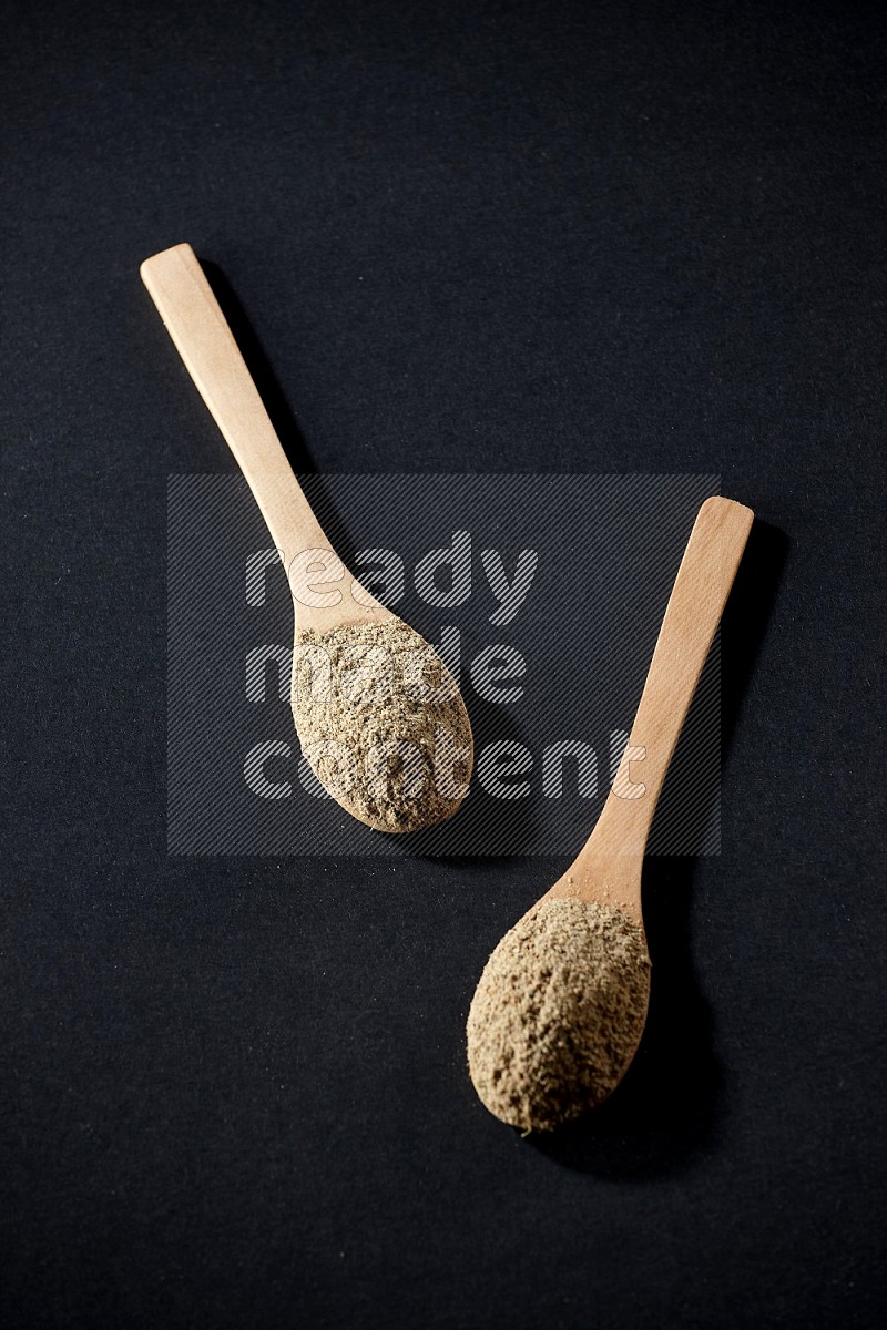 2 Wooden spoons full of cardamom powder on black flooring