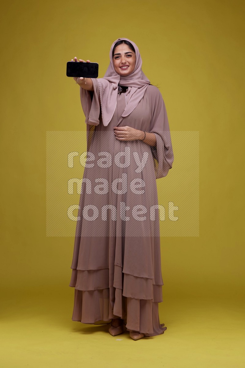 A woman Showing her Phone screen on a Yellow Background wearing Brown Abaya with Hijab