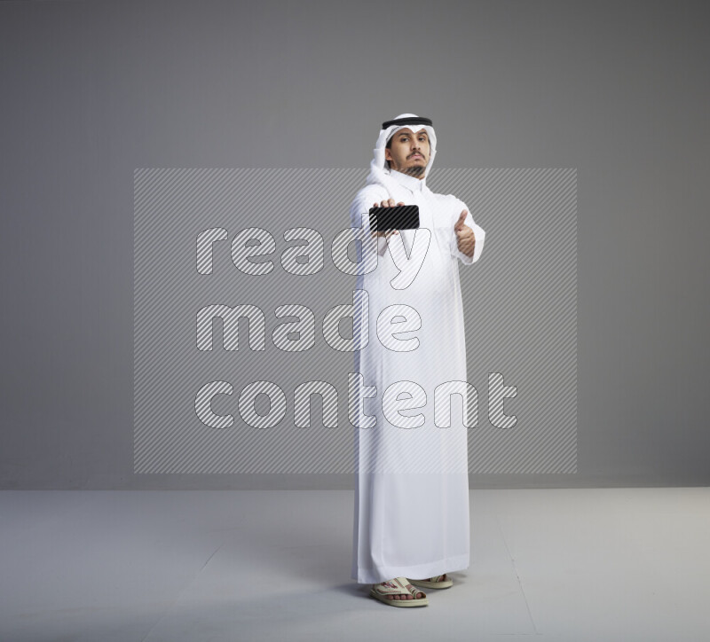 A Saudi man standing wearing thob and white shomag showing phone to camera on gray background