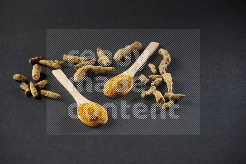 2 wooden spoons full of turmeric powder with dried turmeric fingers on black flooring