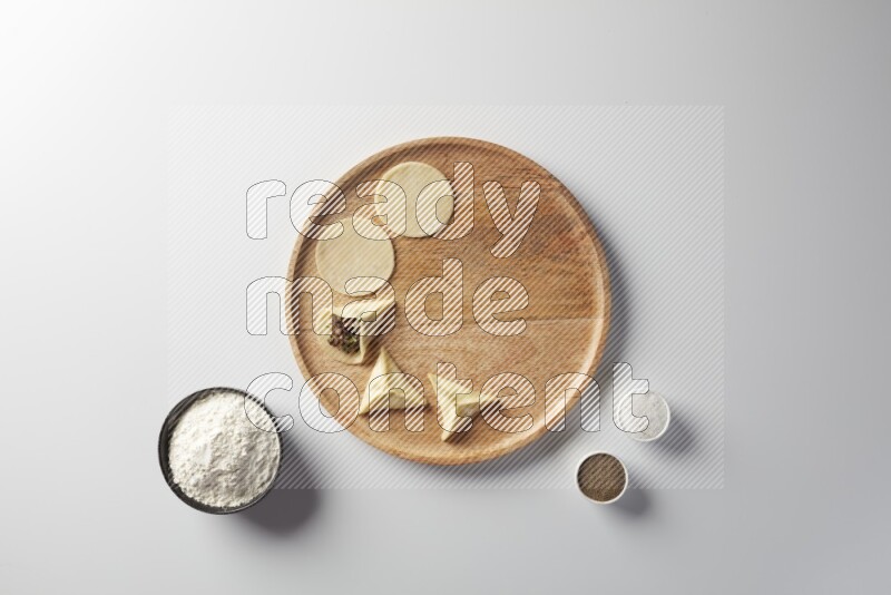 two closed sambosas and one open sambosa filled with meat while flour, salt, and black pepper aside in a wooden dish on a white background