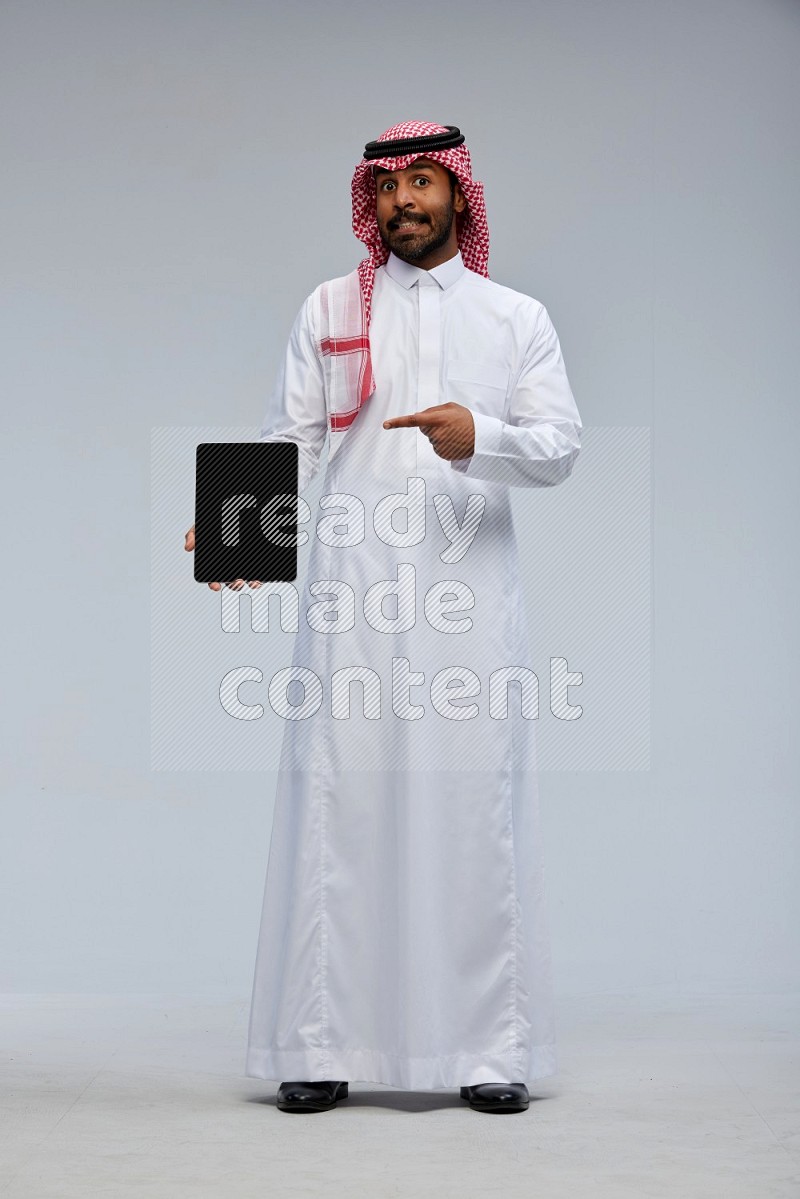 Saudi man Wearing Thob and shomag standing showing tablet to camera on Gray background