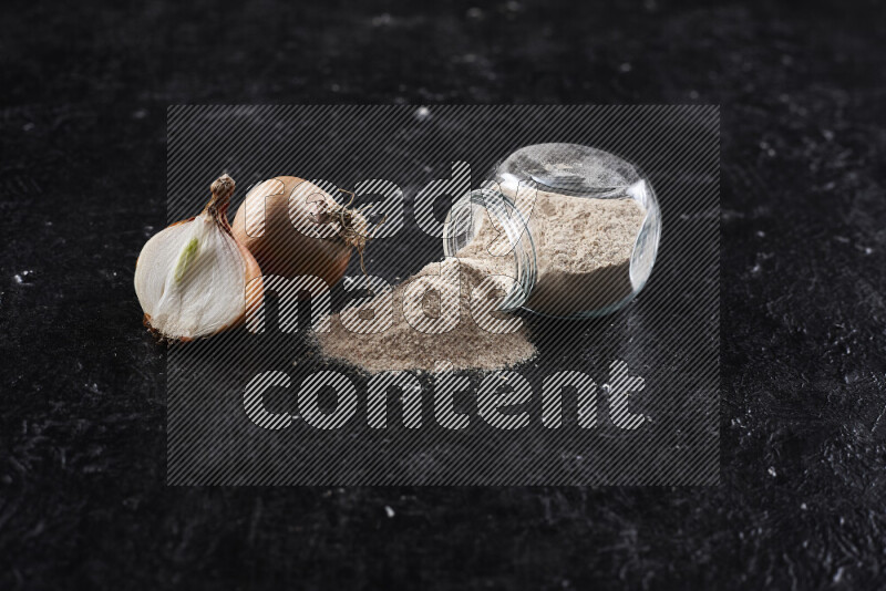 A glass jar full of onion powder flipped with some spilling powder on black background