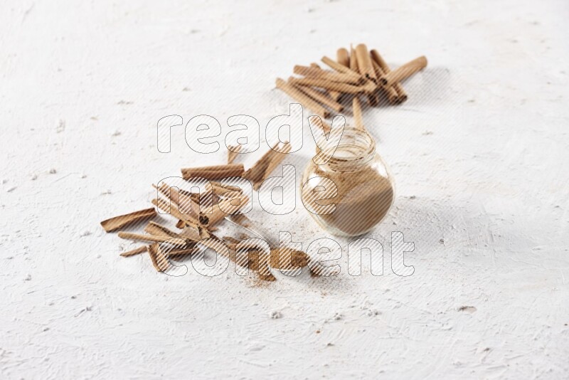 Herbal glass jar full cinnamon powder and a metal spoon surrounded by cinnamon sticks on a white background