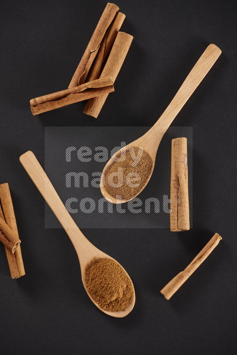 Cinnamon powder in two wooden spoons with cinnamon sticks on black background