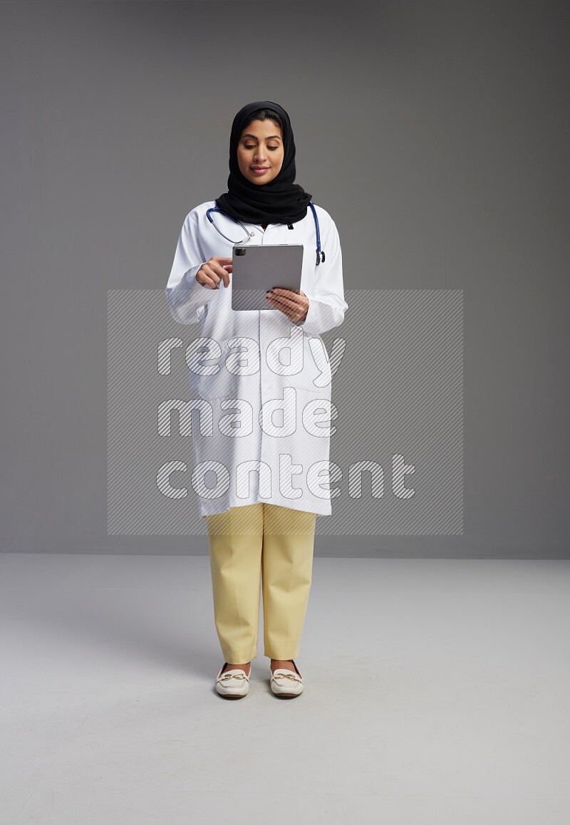 Saudi woman wearing lab coat with stethoscope standing working on tablet on Gray background