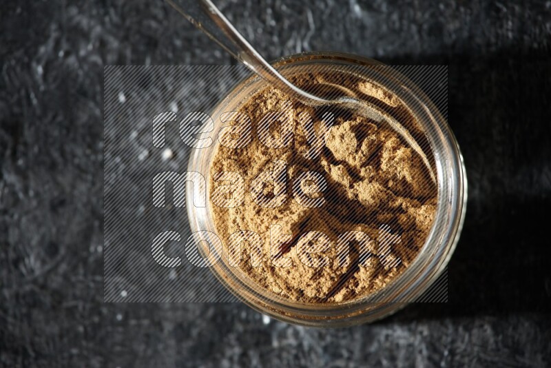 A glass jar and a metal spoon full of allspice powder on a textured black flooring