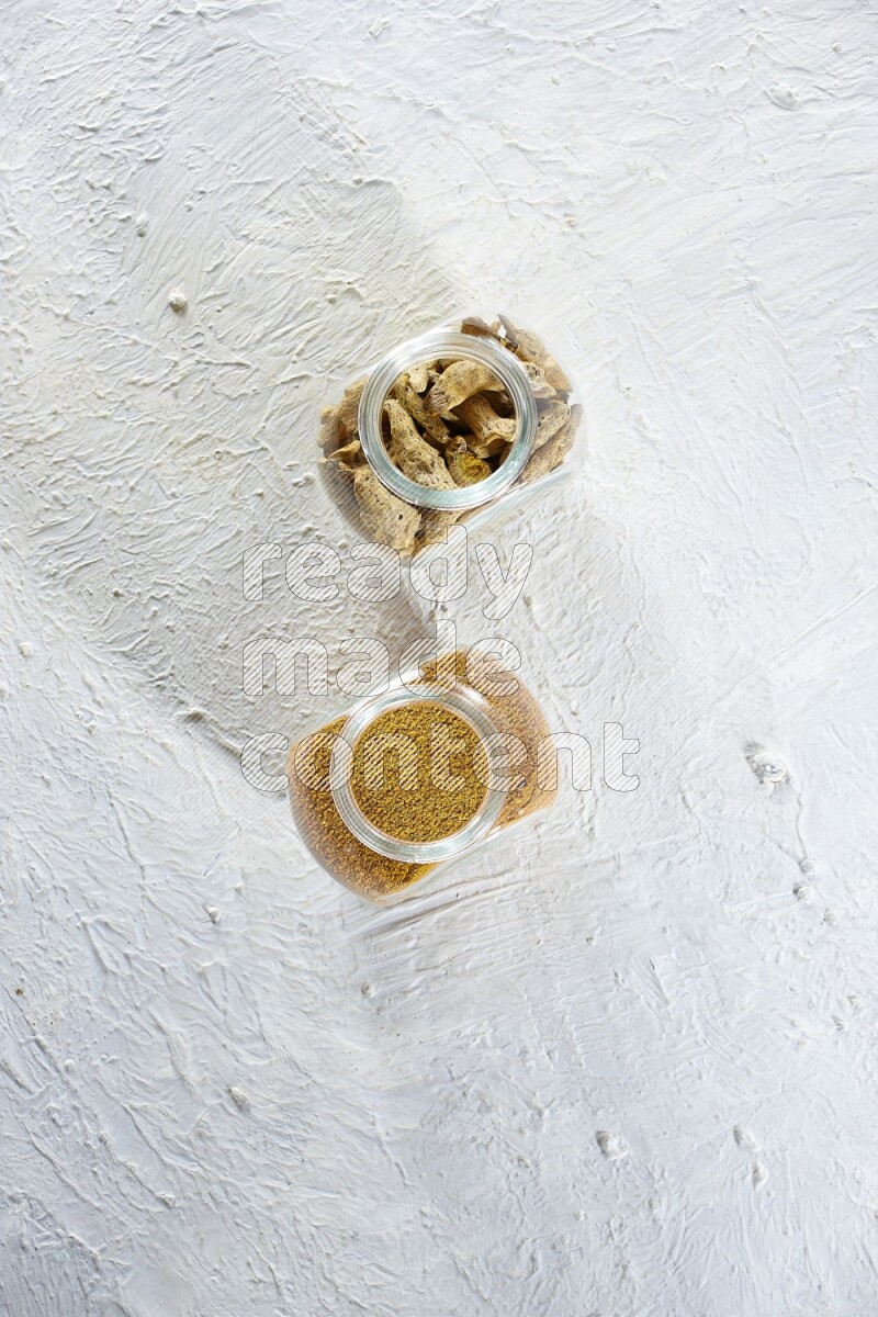 2 glass spice jars full of turmeric powder and dried whole fingers on textured white flooring
