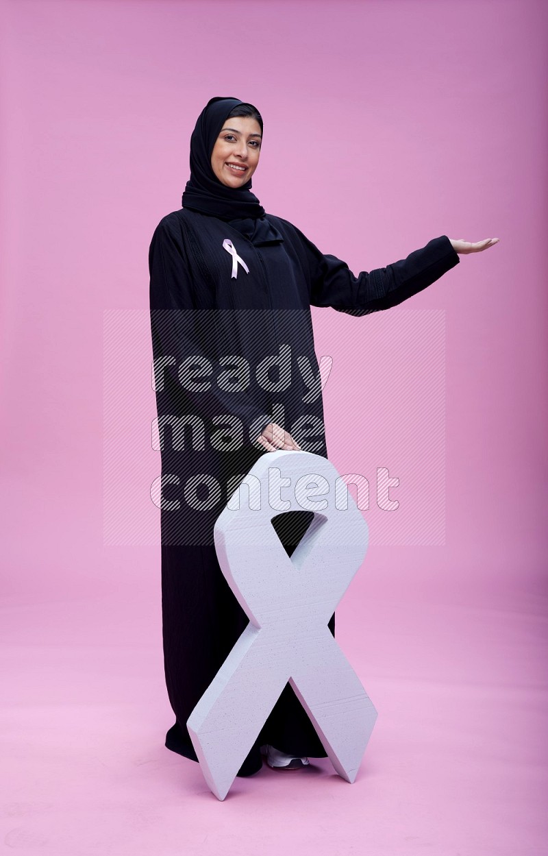 Saudi woman wearing pink ribbon on abaya standing holding awareness ribbon on plain pink background