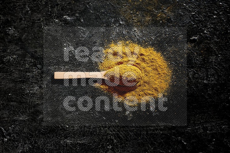 A wooden spoon full of turmeric powder on textured black background