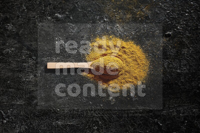 A wooden spoon full of turmeric powder on textured black background