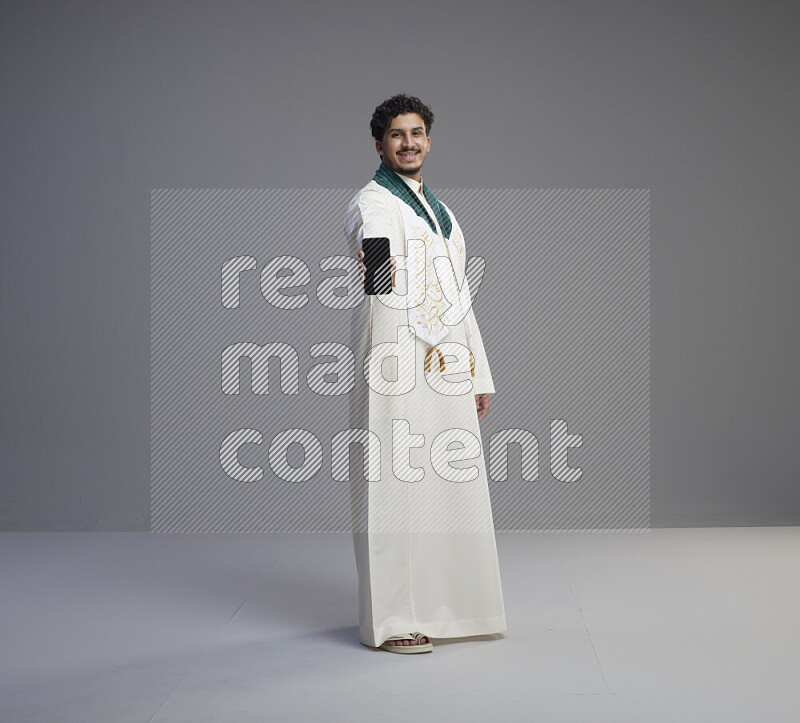 A Saudi man standing wearing thob and Saudi flag scarf showing phone to camera on gray background