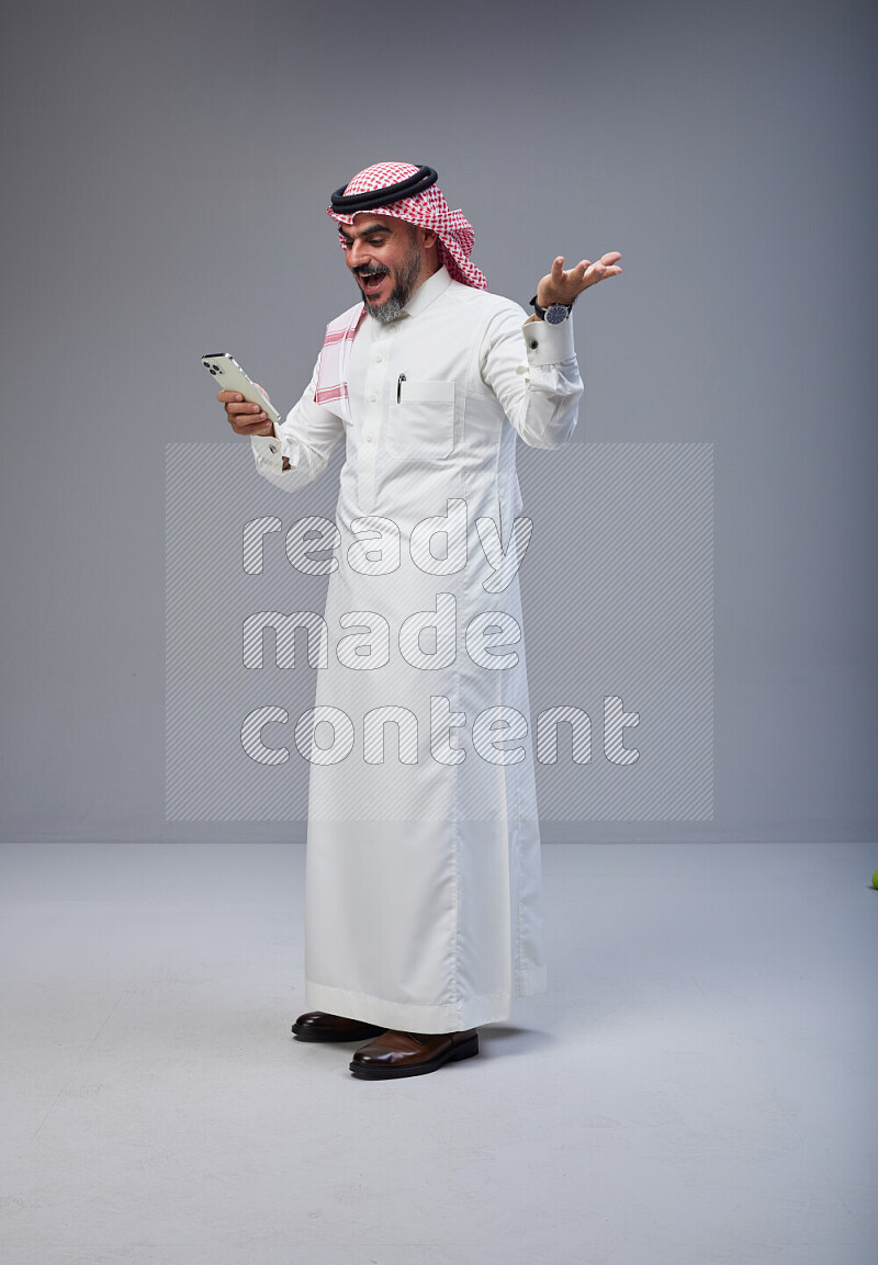 Saudi man Wearing Thob and red Shomag standing texting on phone on Gray background