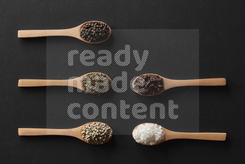 الفلفل الأبيض والقرنفل والهيل والملح والفلفل الأسود وريحان مجفف في معالق خشبية علي خلفية سوداء