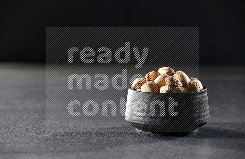 A black pottery bowl full of almonds on a black background in different angles