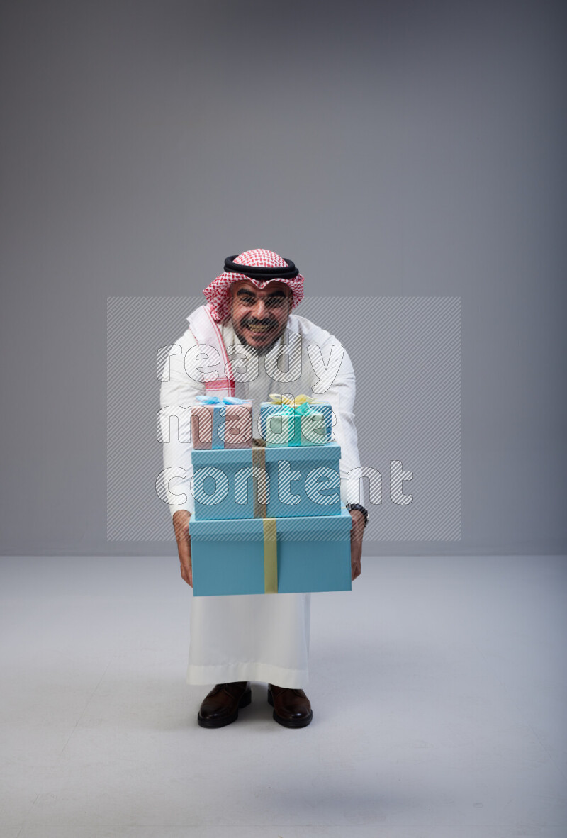 Saudi man Wearing Thob and red Shomag standing holding gift box on Gray background