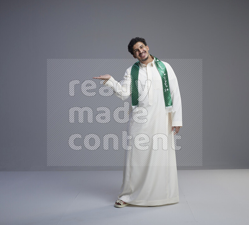 A Saudi man standing wearing thob and Saudi flag scarf on gray background