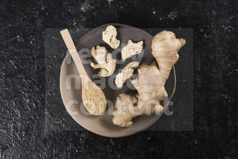 A wooden spoon full of ground ginger powder with fresh and dried ginger, all on a pottery plate on black background