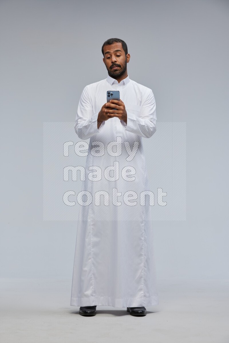 Saudi man wearing Thob standing texting on phone on Gray background