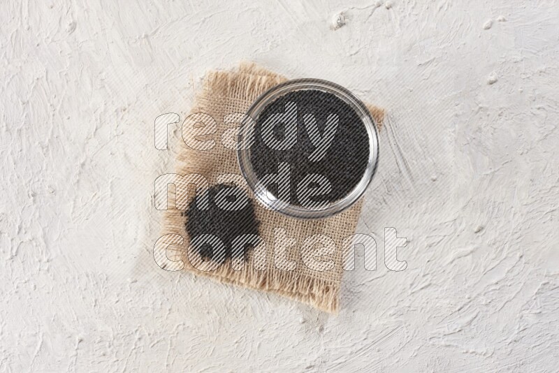 A glass bowl full of black seeds on a burlap piece on textured white flooring