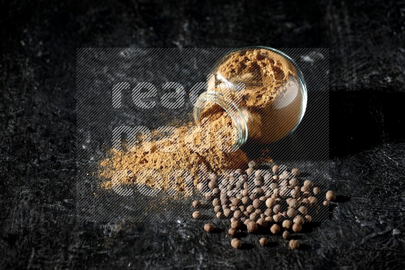 A flipped glass spice jar full of allspice powder and powder spilled out of it with whole balls on a textured black flooring