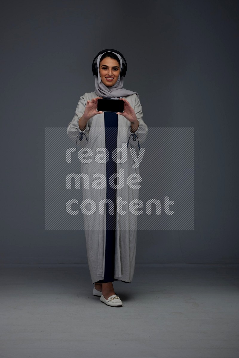 A Saudi woman wearing a light gray Abaya and head scarf setting on a dark grey chair and watching on the tablet eye level on a grey background