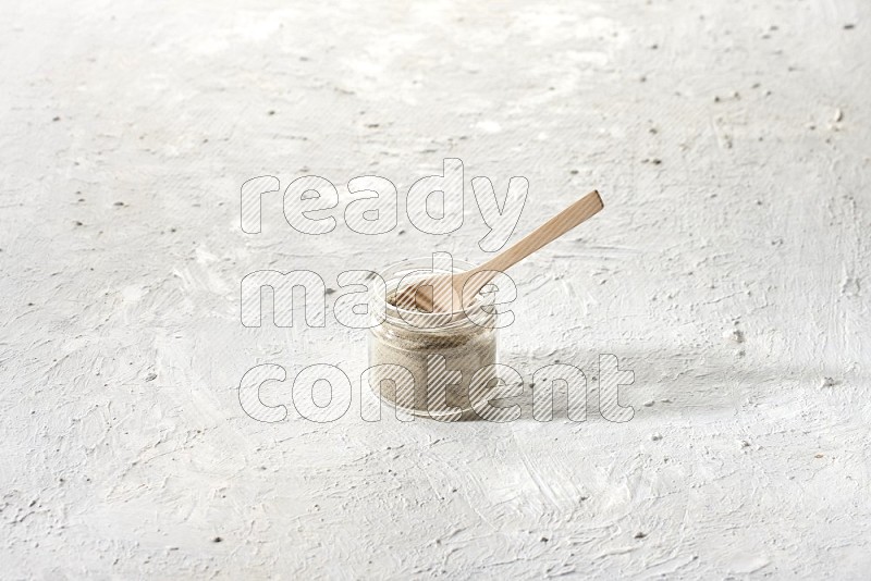 A glass jar and wooden spoon full of white pepper powder on textured white flooring