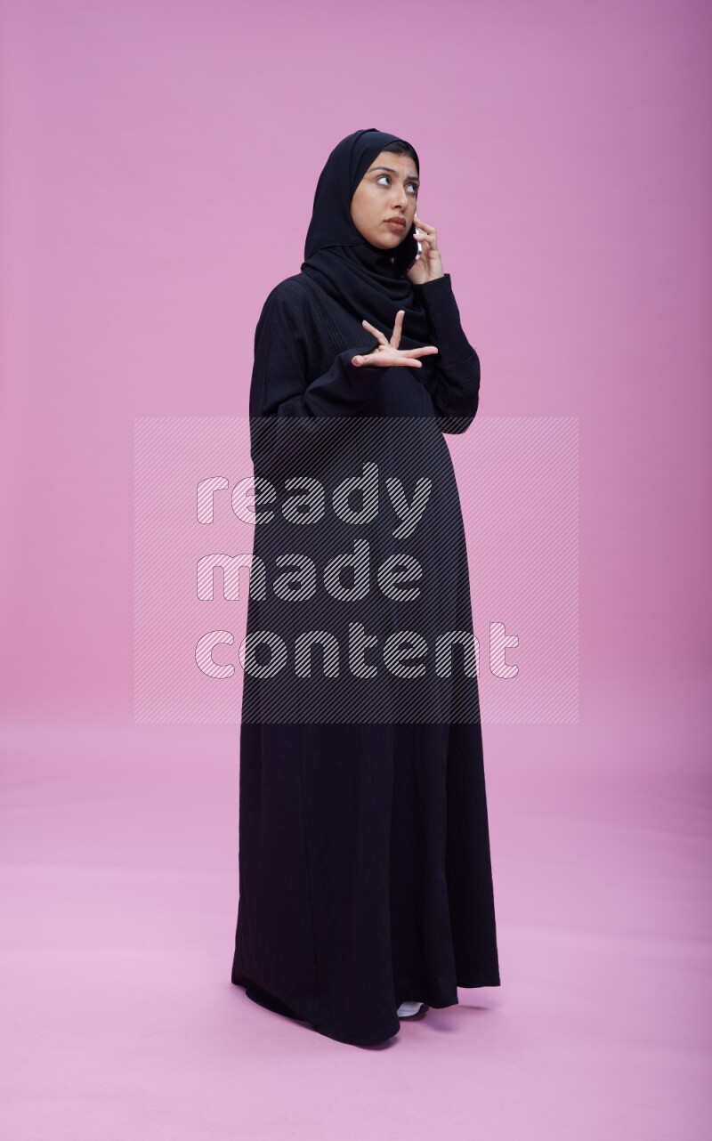 Saudi woman wearing Abaya standing talking on phone on pink background