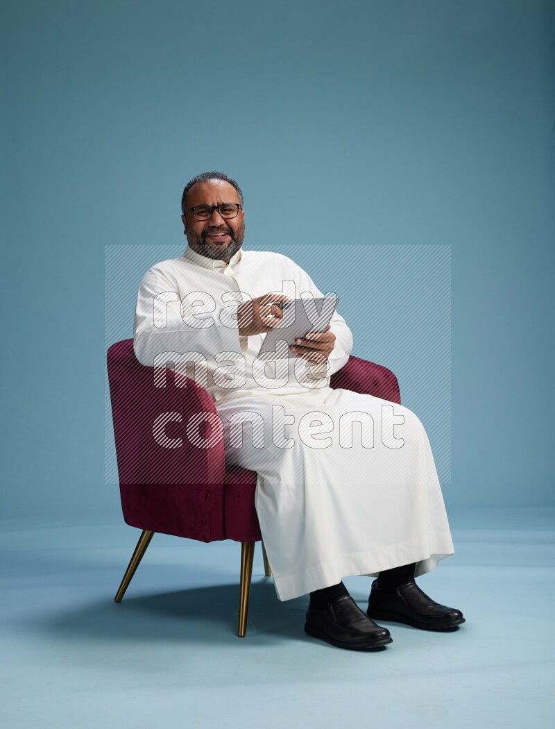 Saudi Man without shimag sitting on chair working on tablet on blue background
