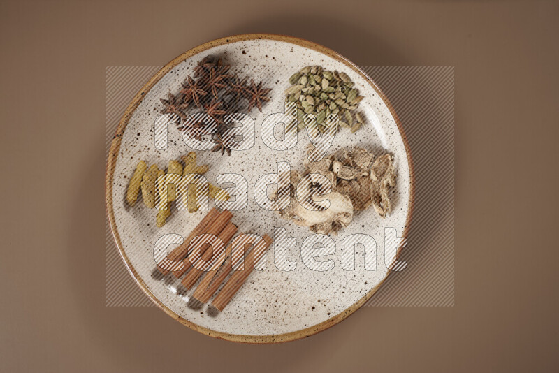 An assorted whole spices on a flat beige plate including fresh and dried ginger, cinnamon sticks, cardamom, turmeric fingers, cloves, star anise on a beige background