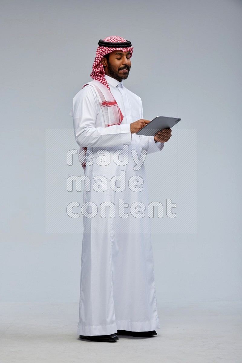 Saudi man Wearing Thob and shomag standing working on tablet on Gray background