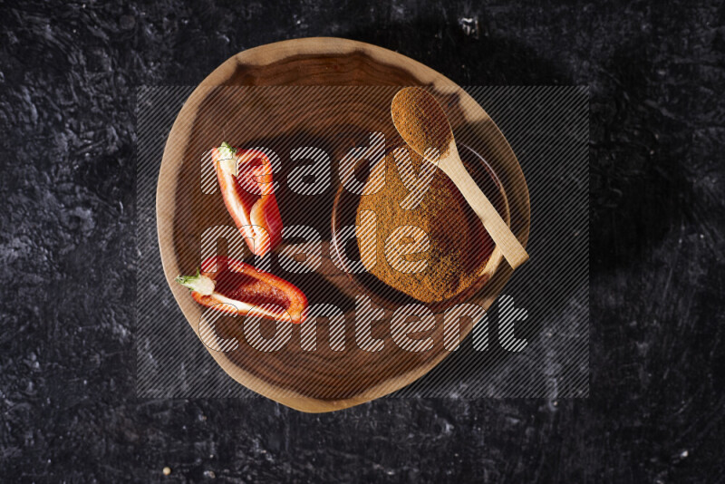 A wooden bowl full of ground paprika powder and sliced red bell pepper beside it, all on a wooden tray on black background