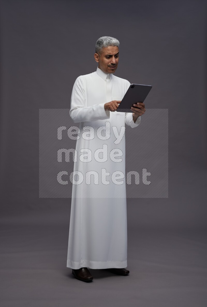 Saudi man wearing thob standing working on tablet on gray background