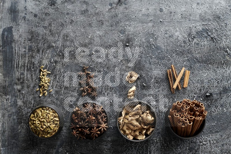Ginger, Cardamom, Star anise and cinnamon sticks in 4 bowls on a textured black background