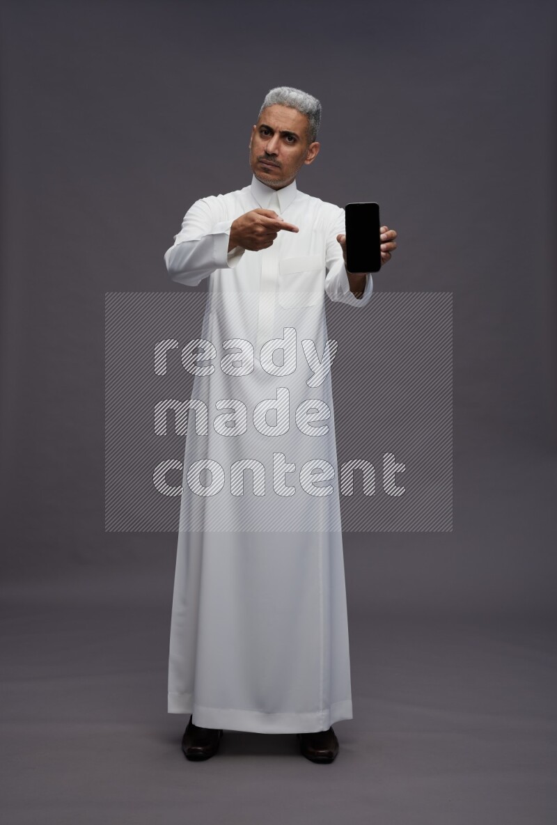 Saudi man wearing thob standing showing phone to camera on gray background