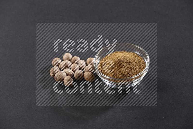 A glass bowl full nutmeg powder with the seeds beside it on a black flooring in different angles