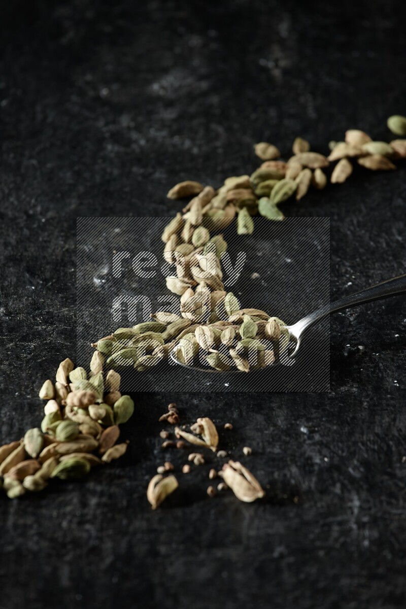 A Metal spoon full of cardamom seeds and some seeds beside it on a textured black flooring