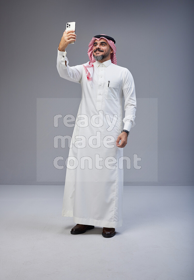 Saudi man Wearing Thob and red Shomag standing taking selfie on Gray background