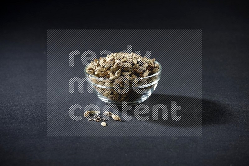 A glass bowl full of cardamom and more seeds spreaded beneath the bowl on black flooring