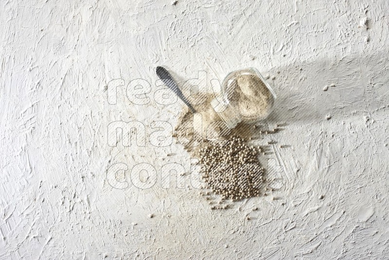 A flipped herbal glass jar and metal spoon full of white pepper powder with spilled powder and beads on textured white flooring
