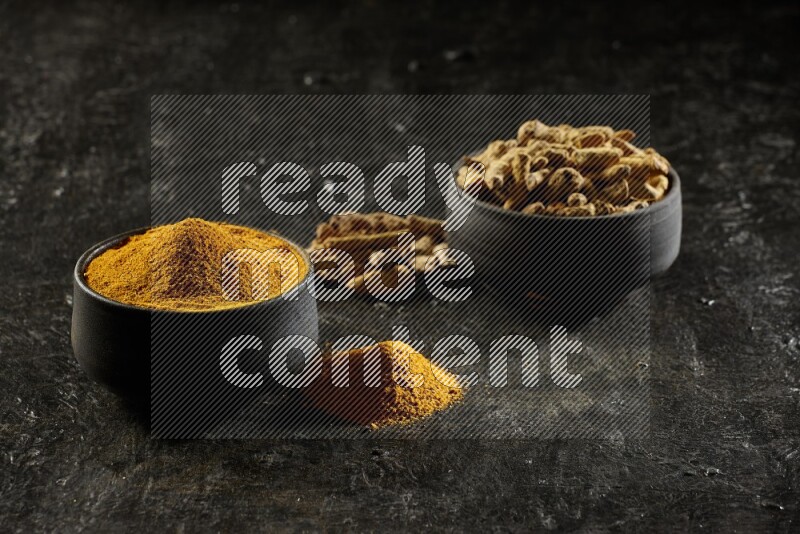2 pottery black bowls, one full of turmeric powder and the other full of dried turmeric whole fingers on a textured black flooring