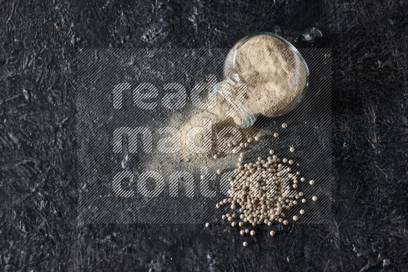 A flipped herbal glass jar full of white pepper powder with spilled powder and pepper beads on textured black flooring
