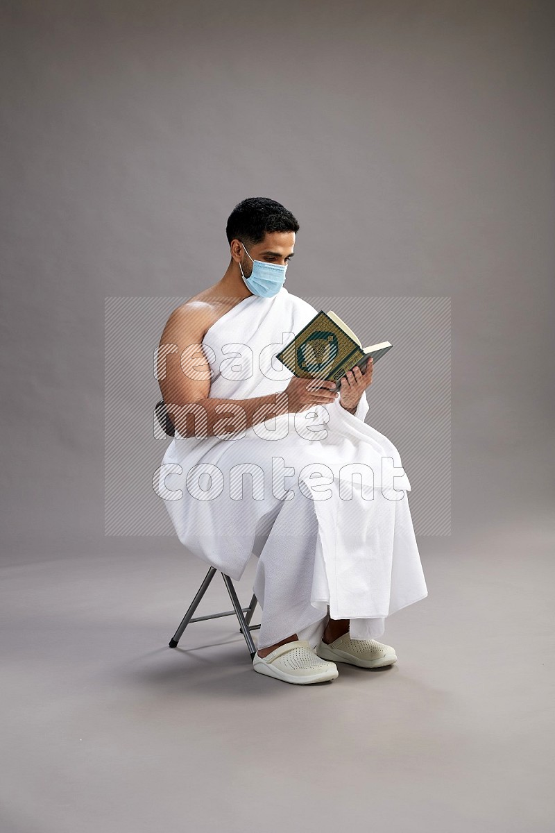 A man wearing Ehram with face mask sitting on chair reading quran on gray background