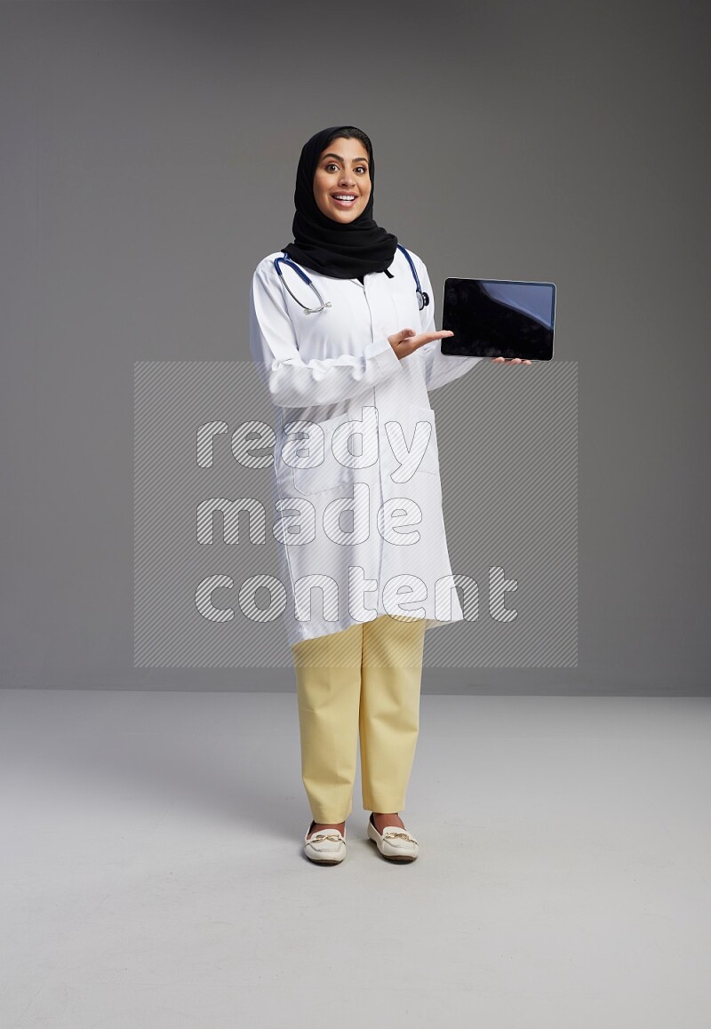 Saudi woman wearing lab coat with stethoscope standing showing tablet to camera with sign in the back on Gray background