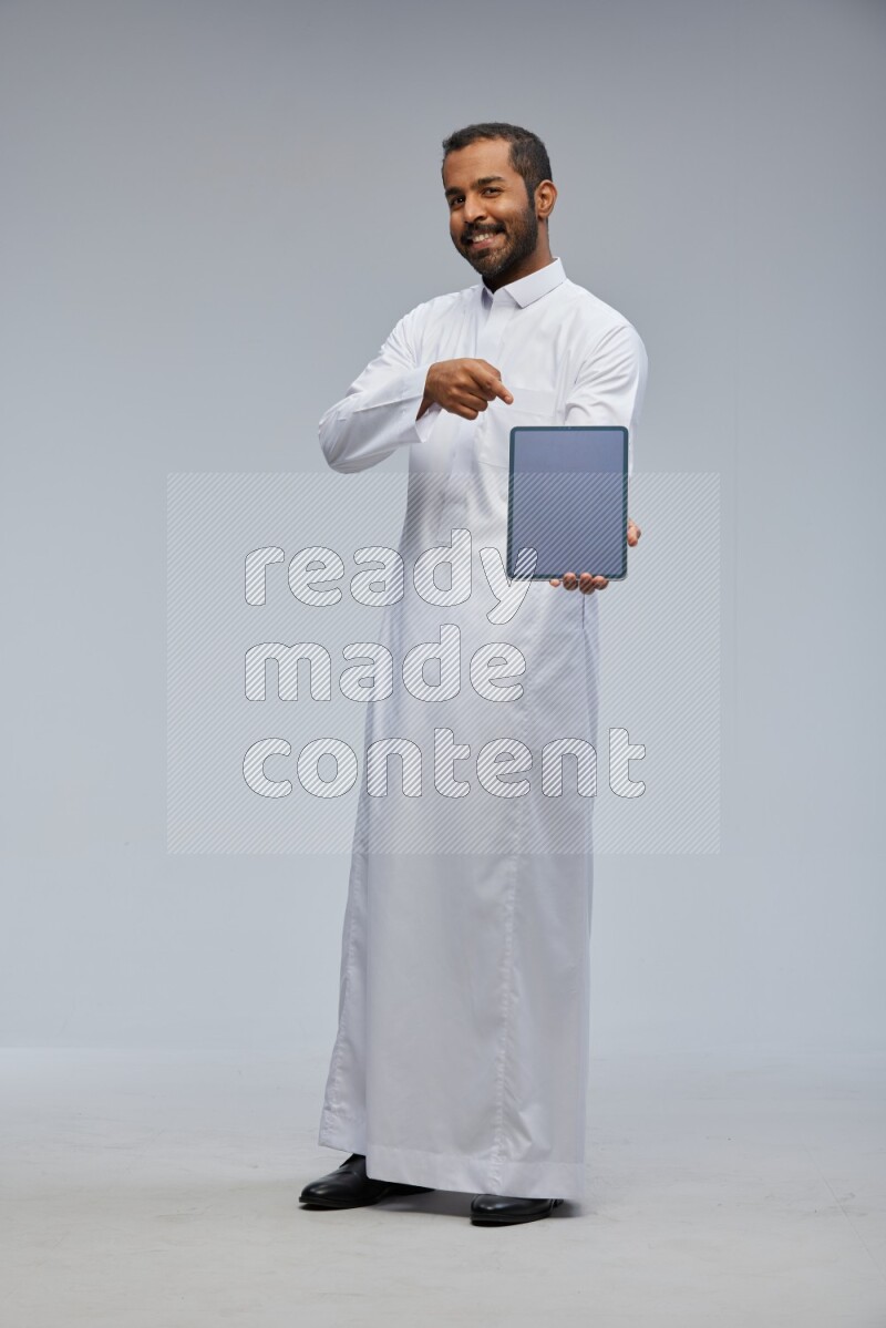 Saudi man wearing Thob standing showing tablet to camera on Gray background